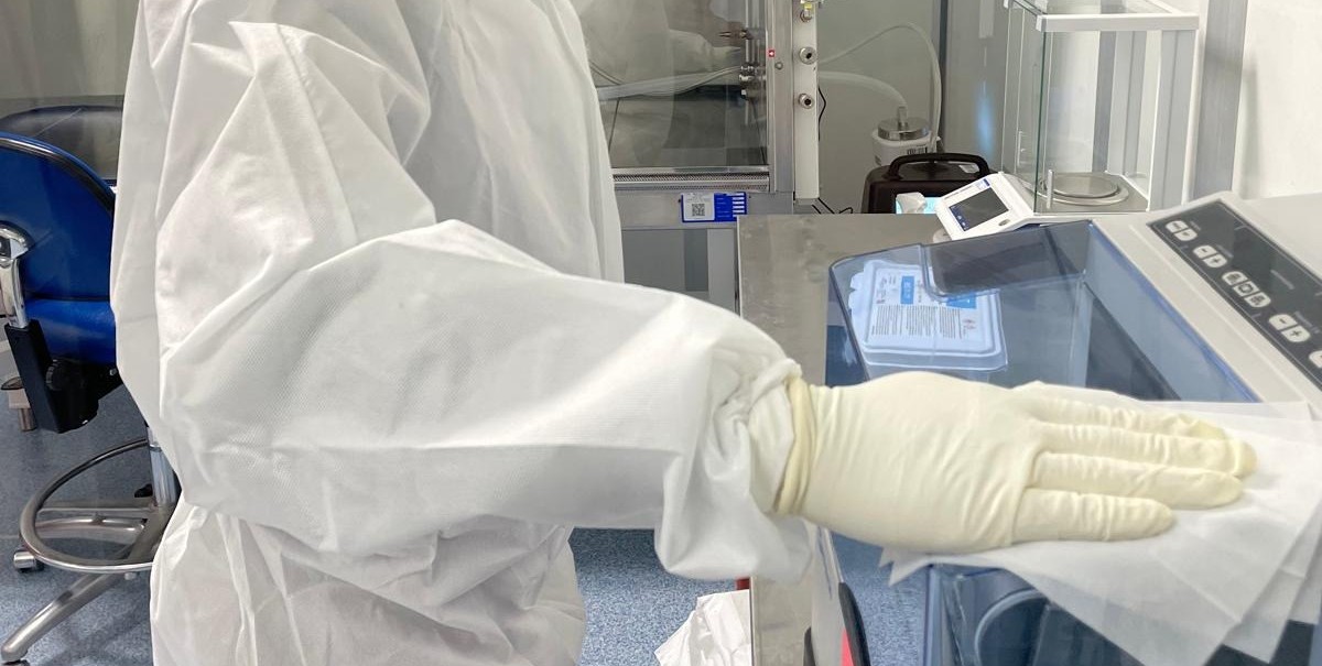 Close view of a gowned arm and a gloved hand drawing a folded wipe across the surface of a piece of equipment, inside a cleanroom.